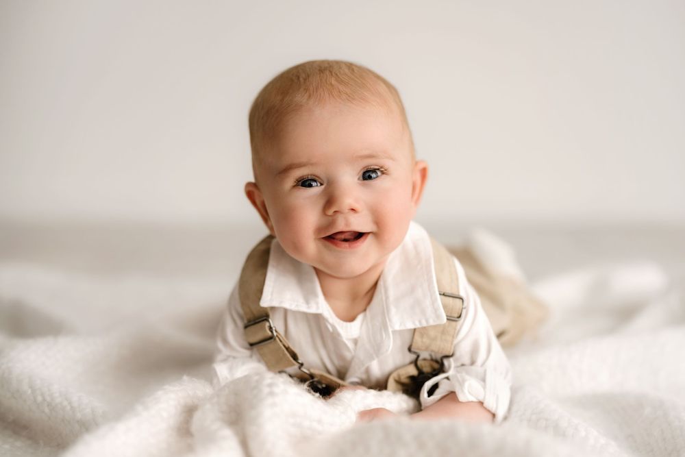 Smiling baby wearing white shirt and beige overalls sitting on soft white blanket.