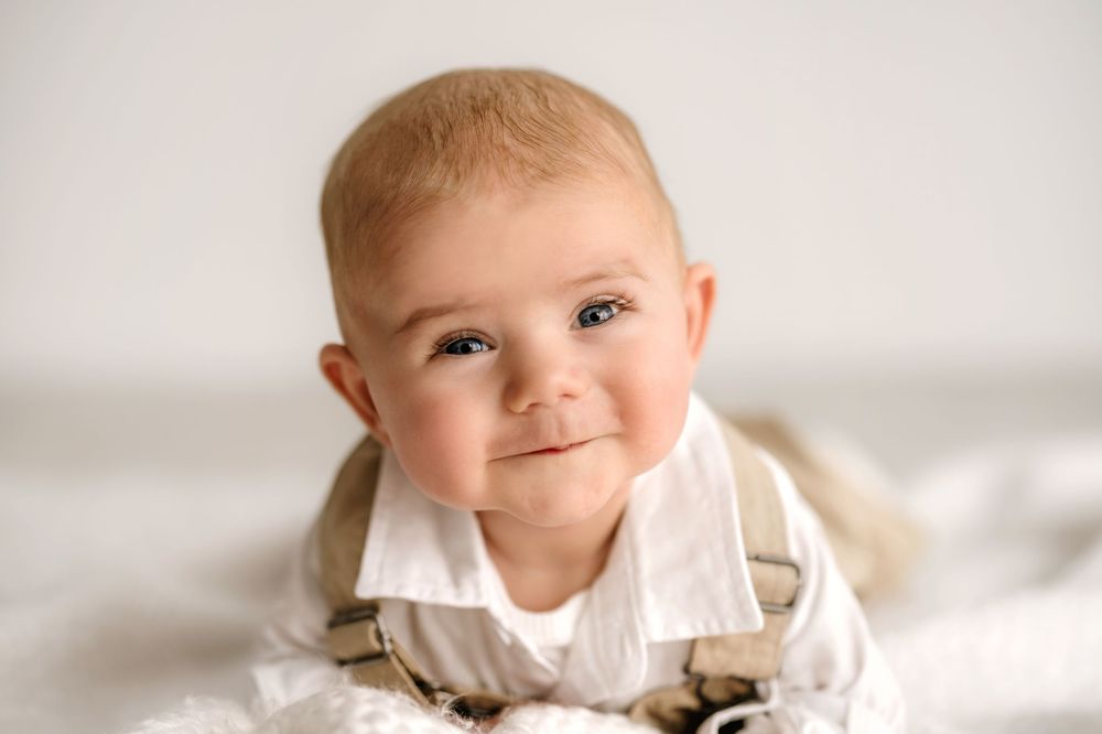 A smiling baby in beige overalls lies on soft white fabric in a portrait studio setting.