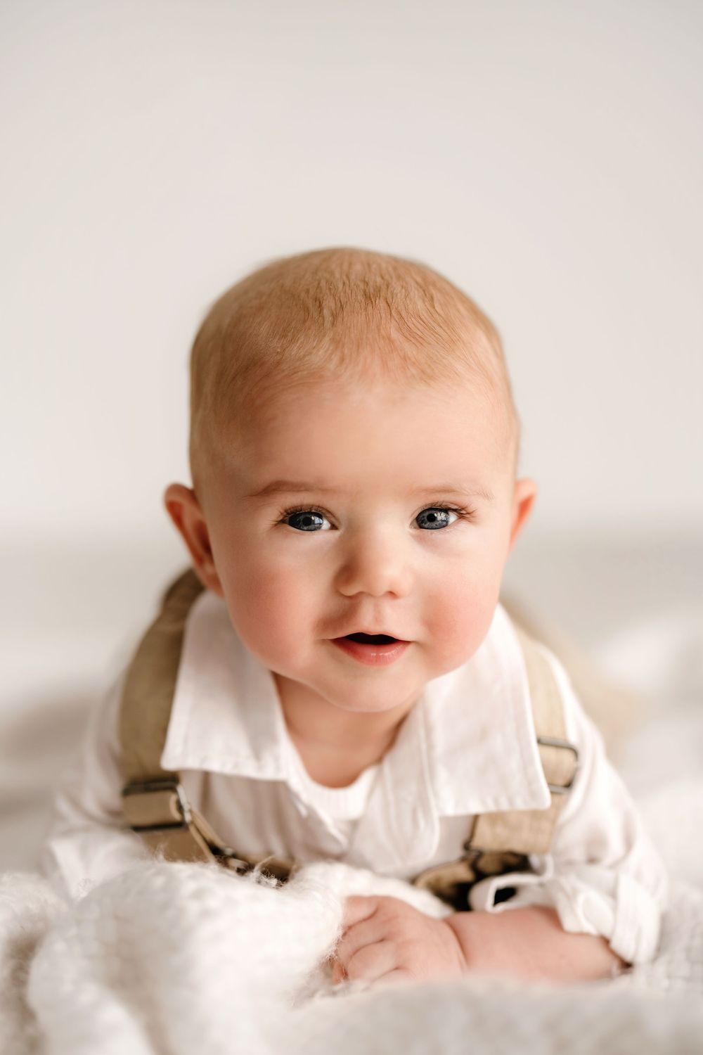 Baby wearing white shirt and beige suspenders lying on soft white blanket with sweet expression.
