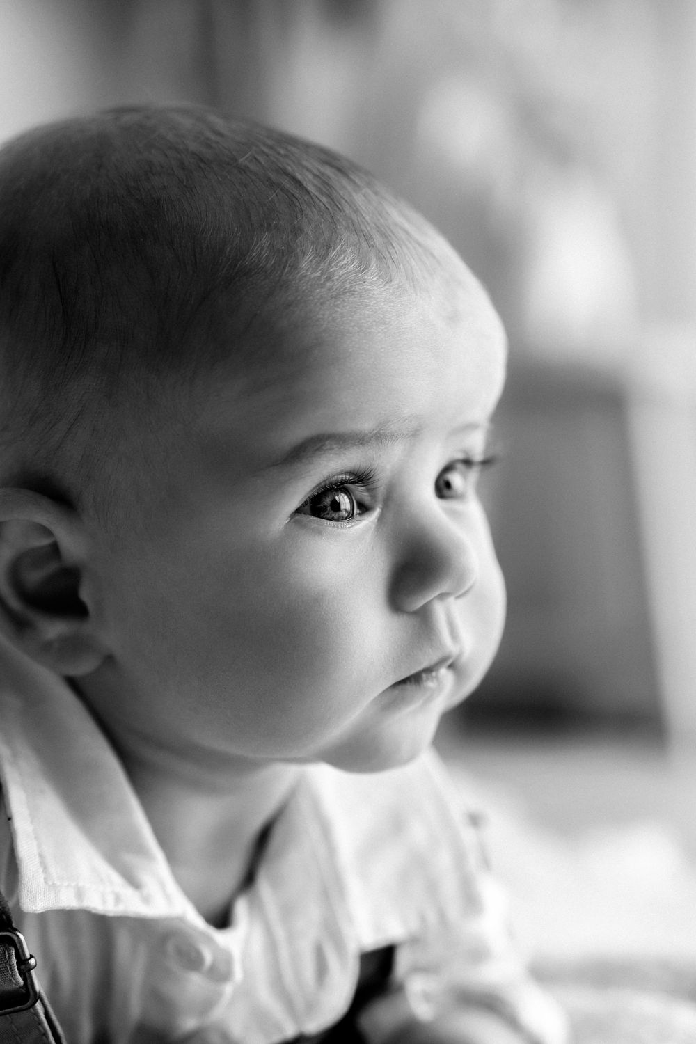 Black and white portrait of a young baby in white clothing looking up with curious expression.