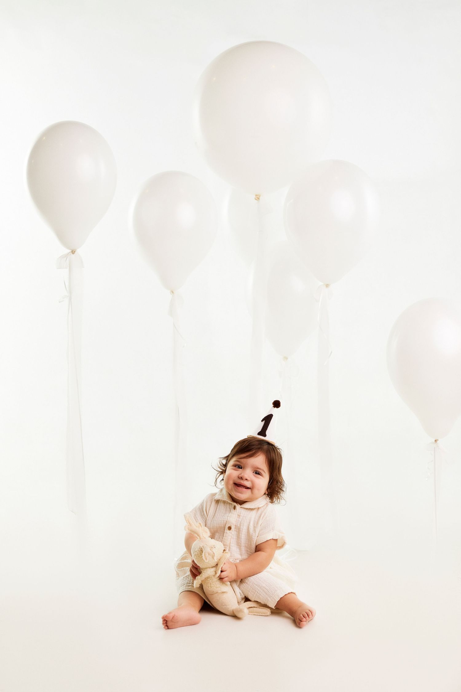 Baby wearing cream outfit and party hat sits among white balloons.
