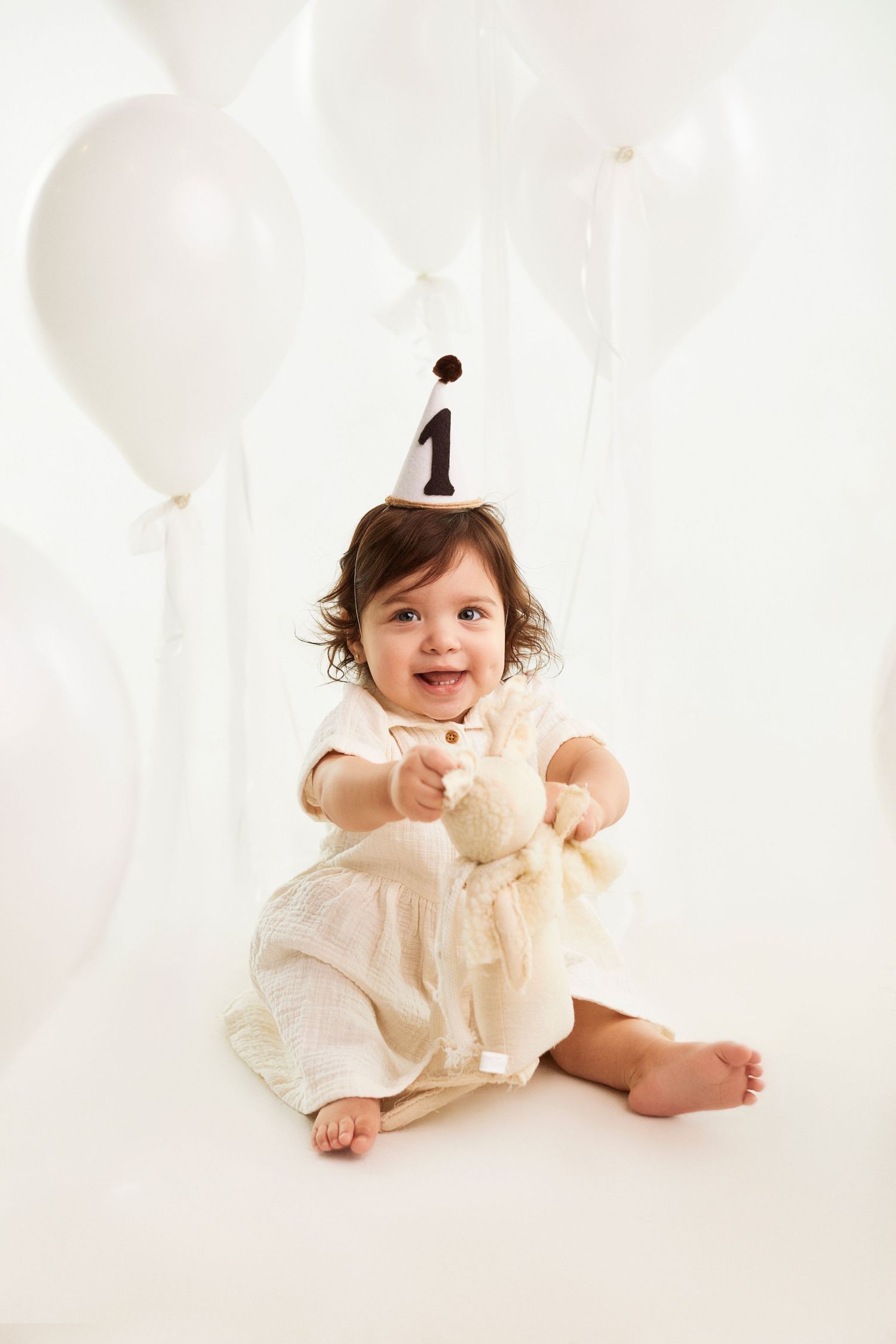 Baby in cream dress with number one hat sits surrounded by white balloons.