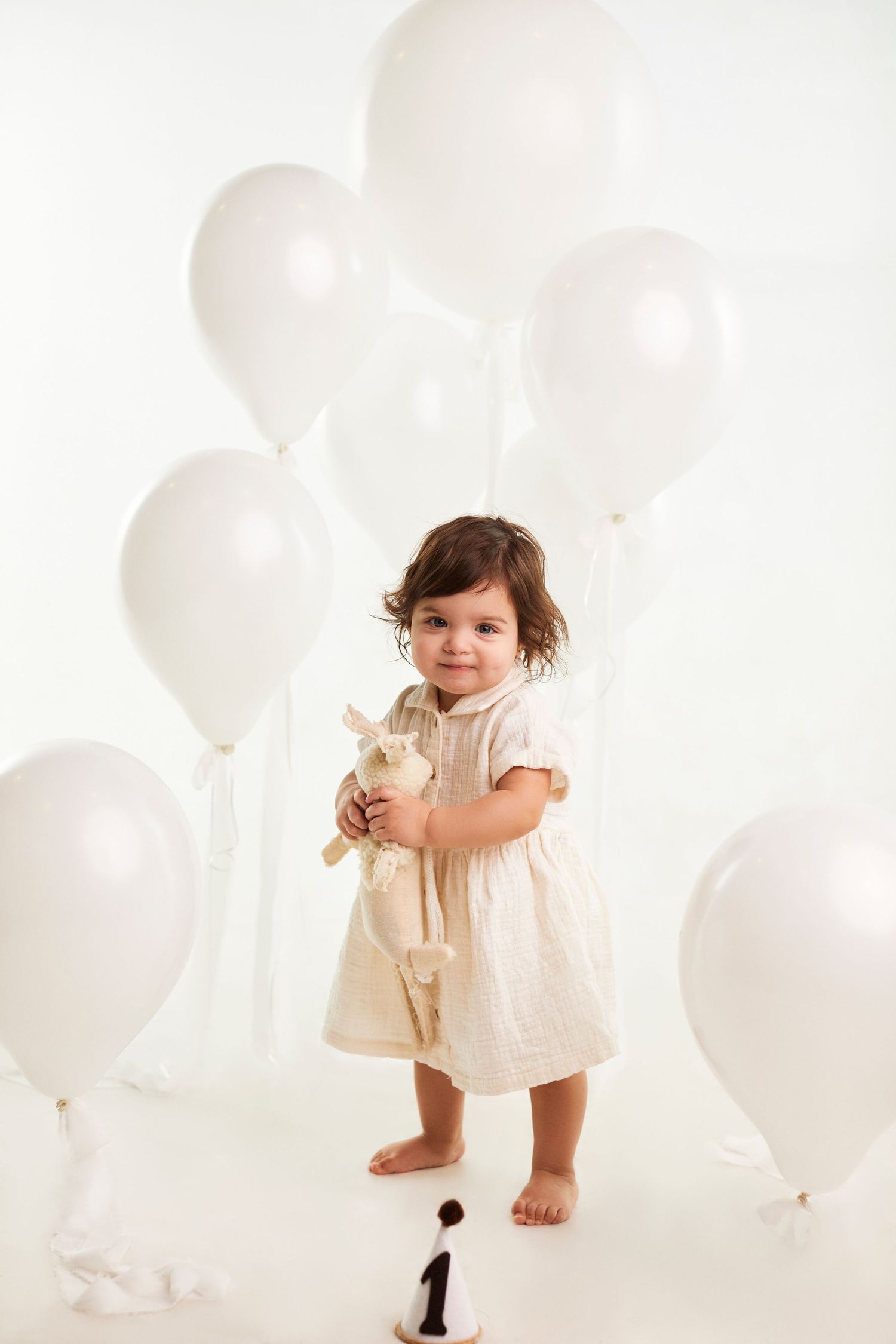 Toddler in cream dress holding stuffed toy among white balloons.