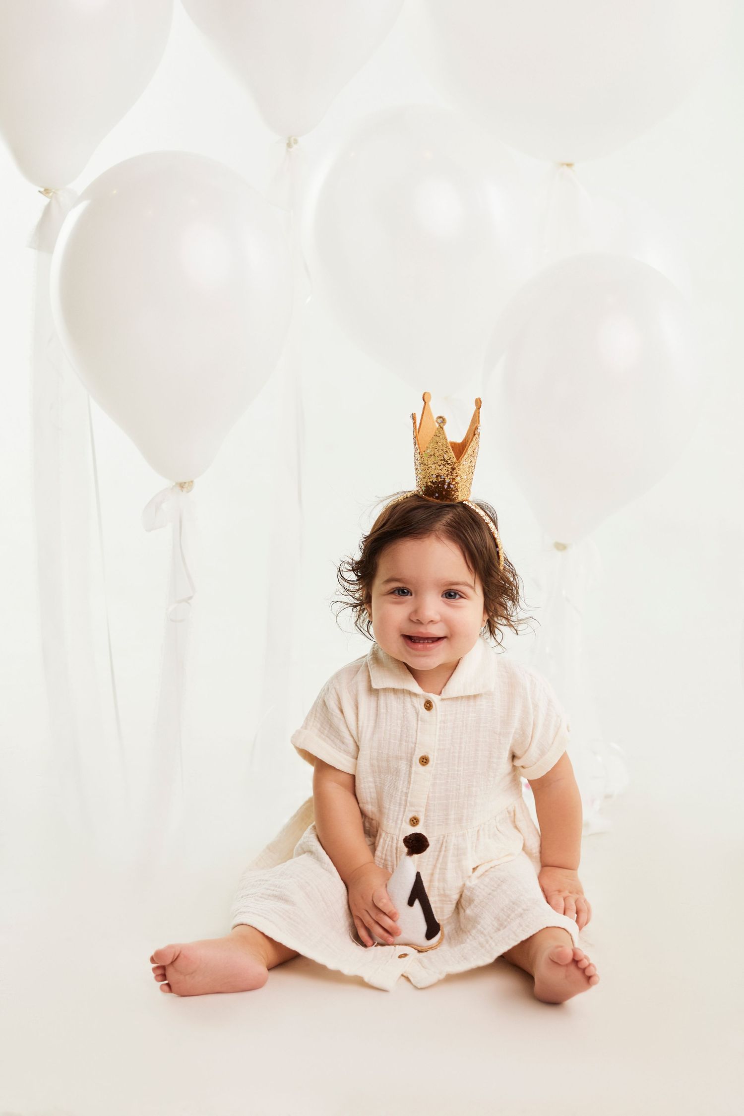 Small child wearing cream dress and gold crown sits among white balloons.