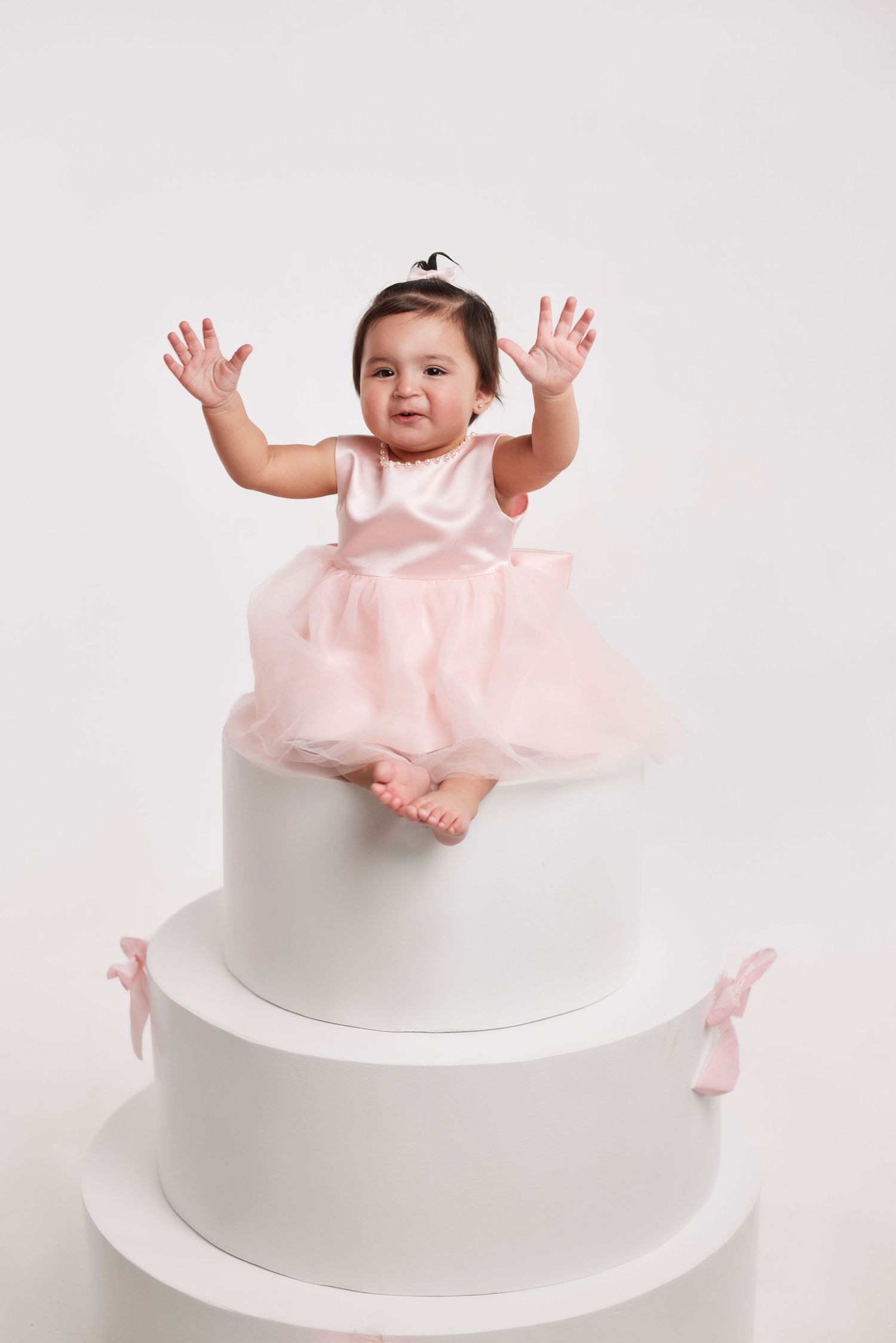 Baby in pink dress playing on white tiered platform during studio photoshoot.