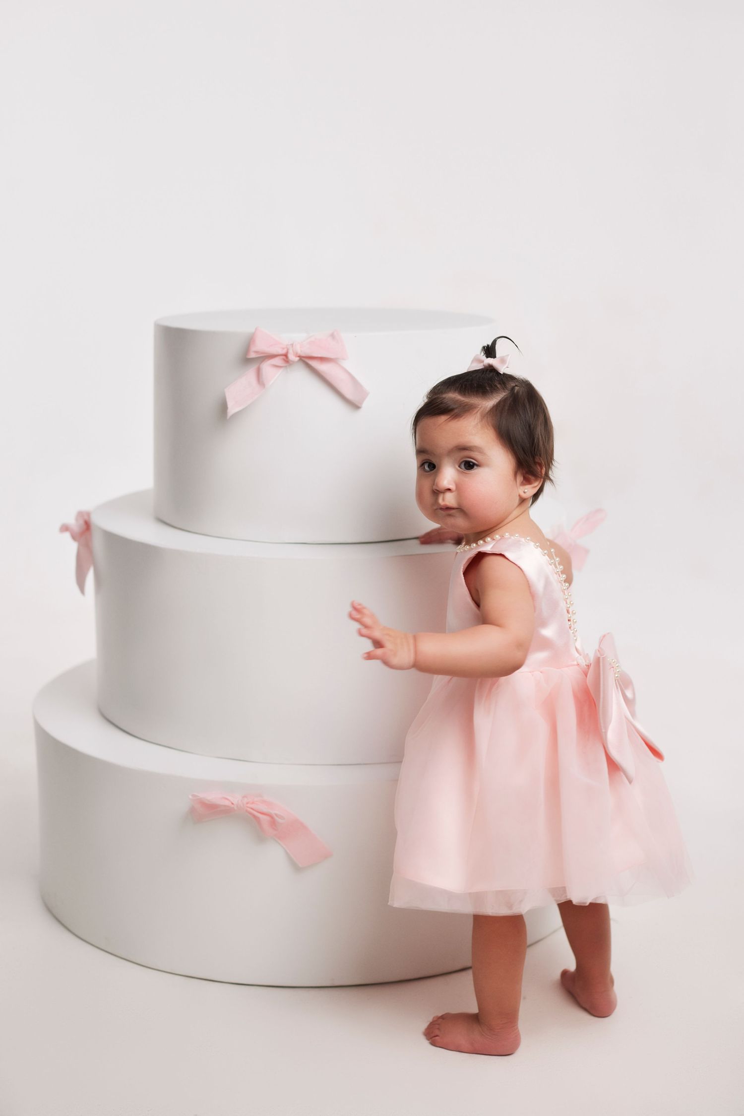 Baby exploring white tiered platform while wearing pink dress during studio session.