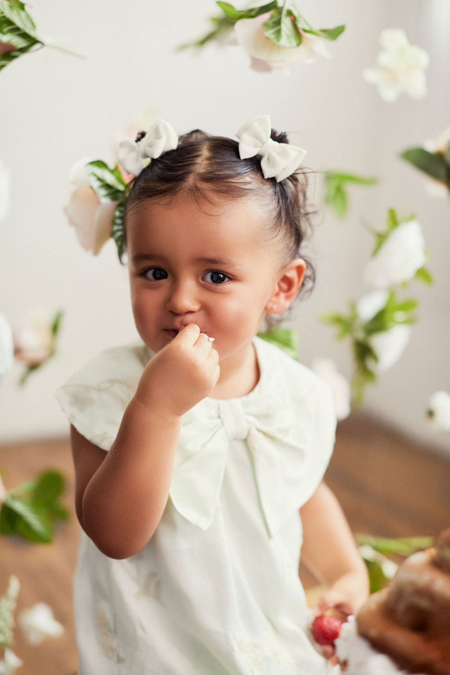 Sweet toddler in white dress with floral hair accessories smiles while playing with flowers.