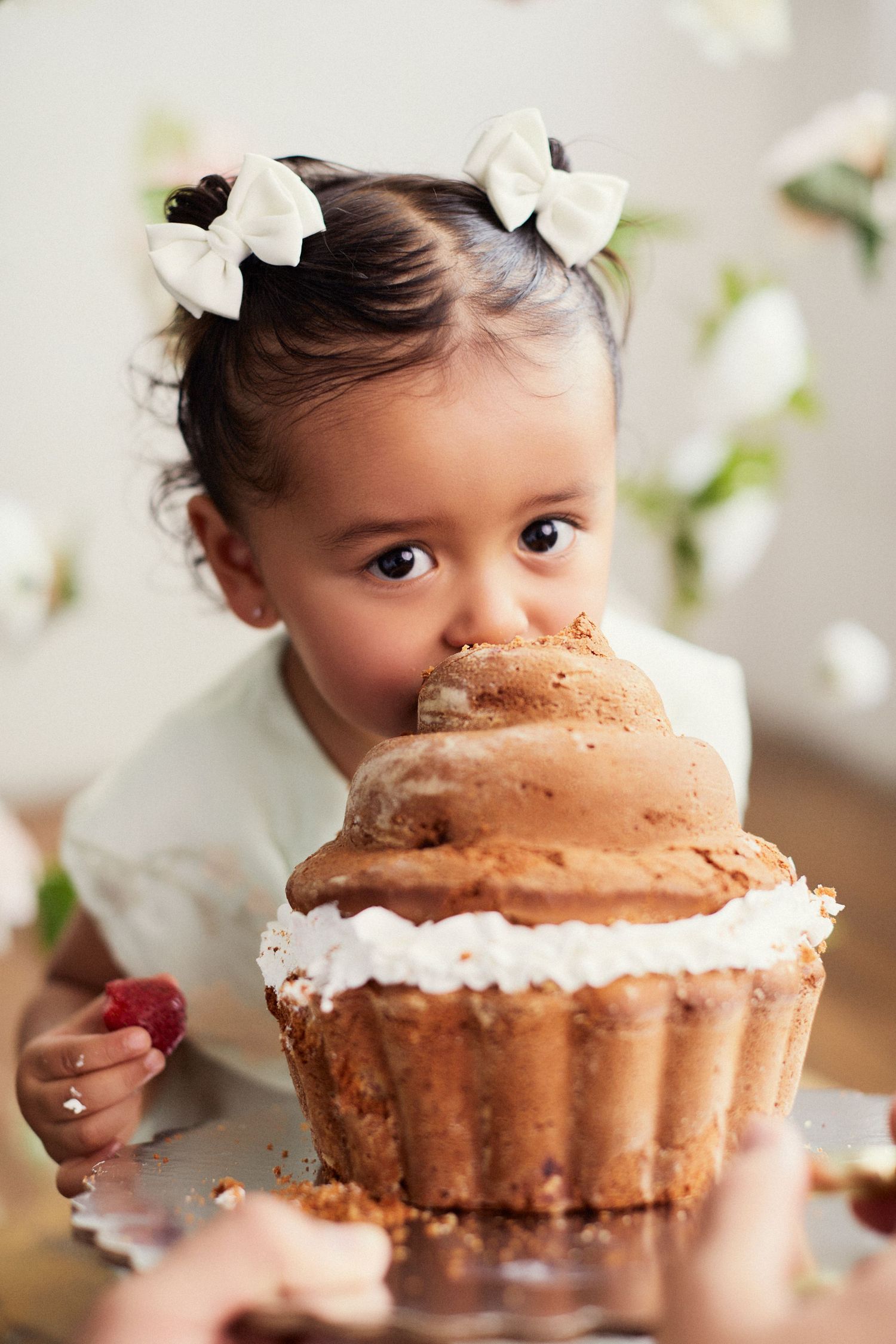 Toddler in white dress enjoying a large chocolate cupcake with white frosting.