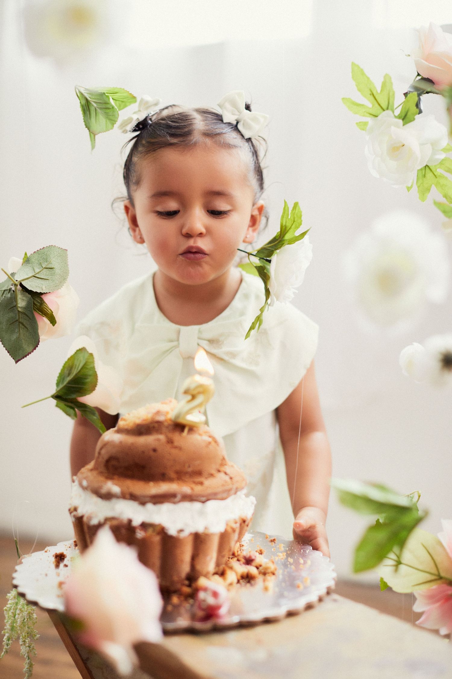 Small child in white outfit blowing on a chocolate cupcake surrounded by white flowers.