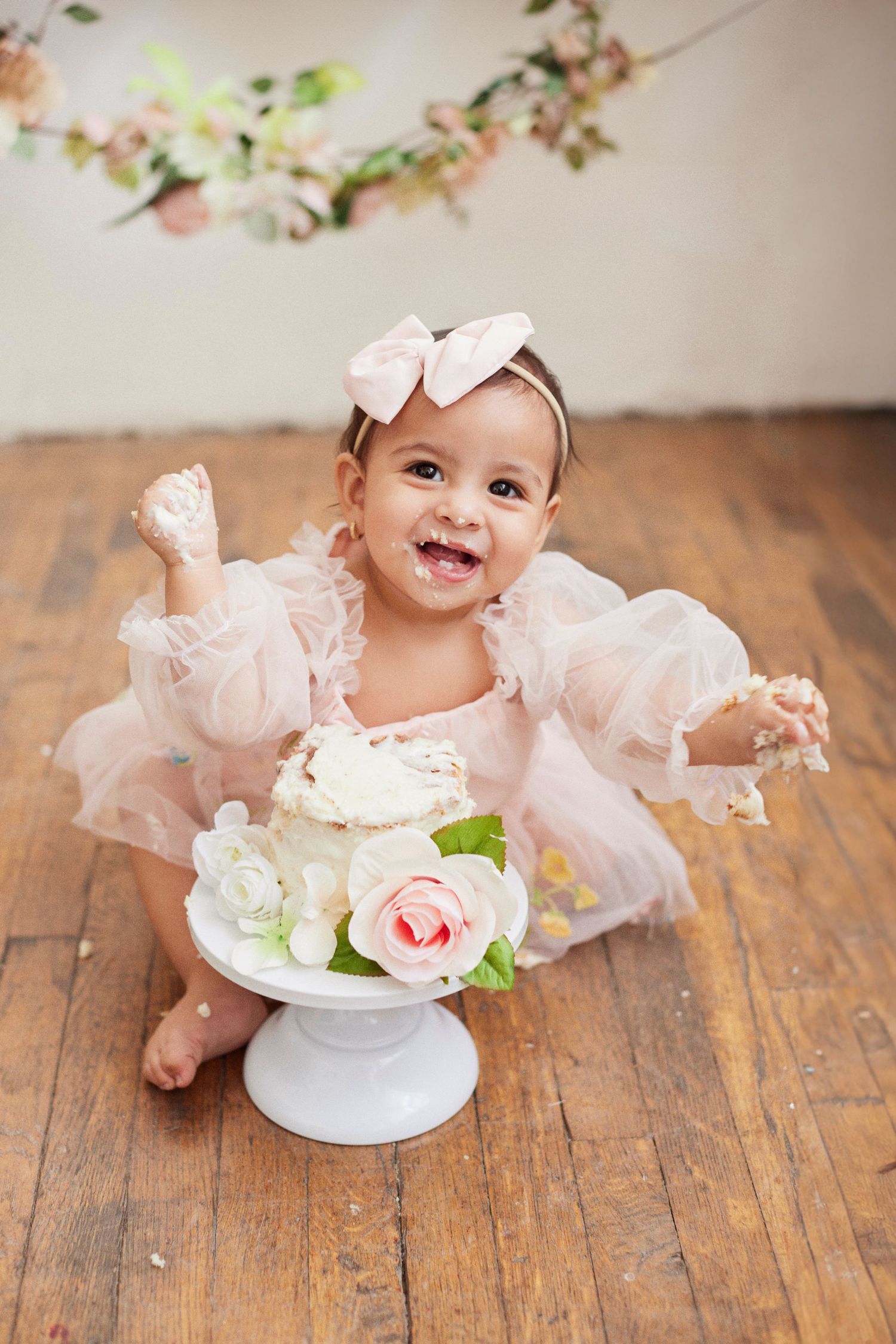 Happy baby enjoying cake smash photography session with floral decorations on wooden floor.
