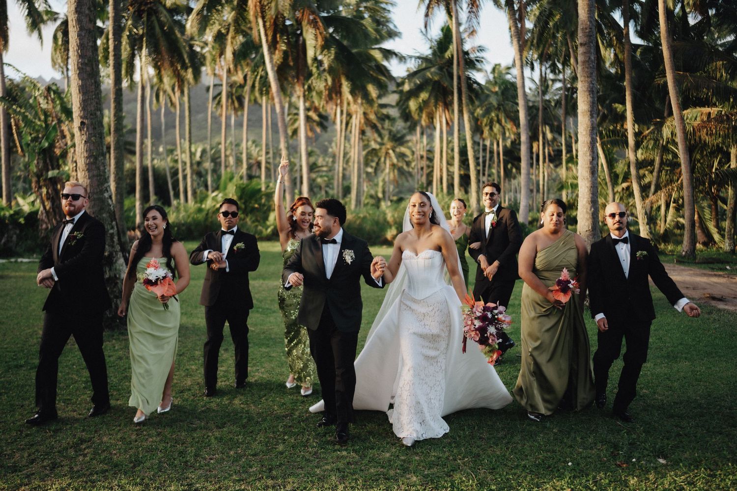 A couple in wedding attire embrace outdoors with tropical palm trees and lush greenery in the background.