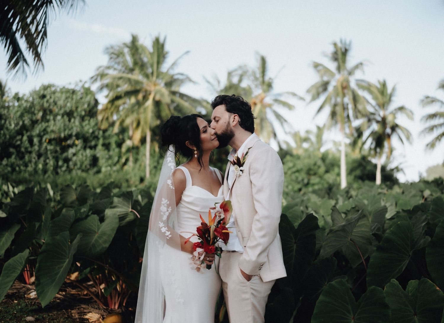 A couple in wedding attire embrace outdoors with tropical palm trees and lush greenery in the background.