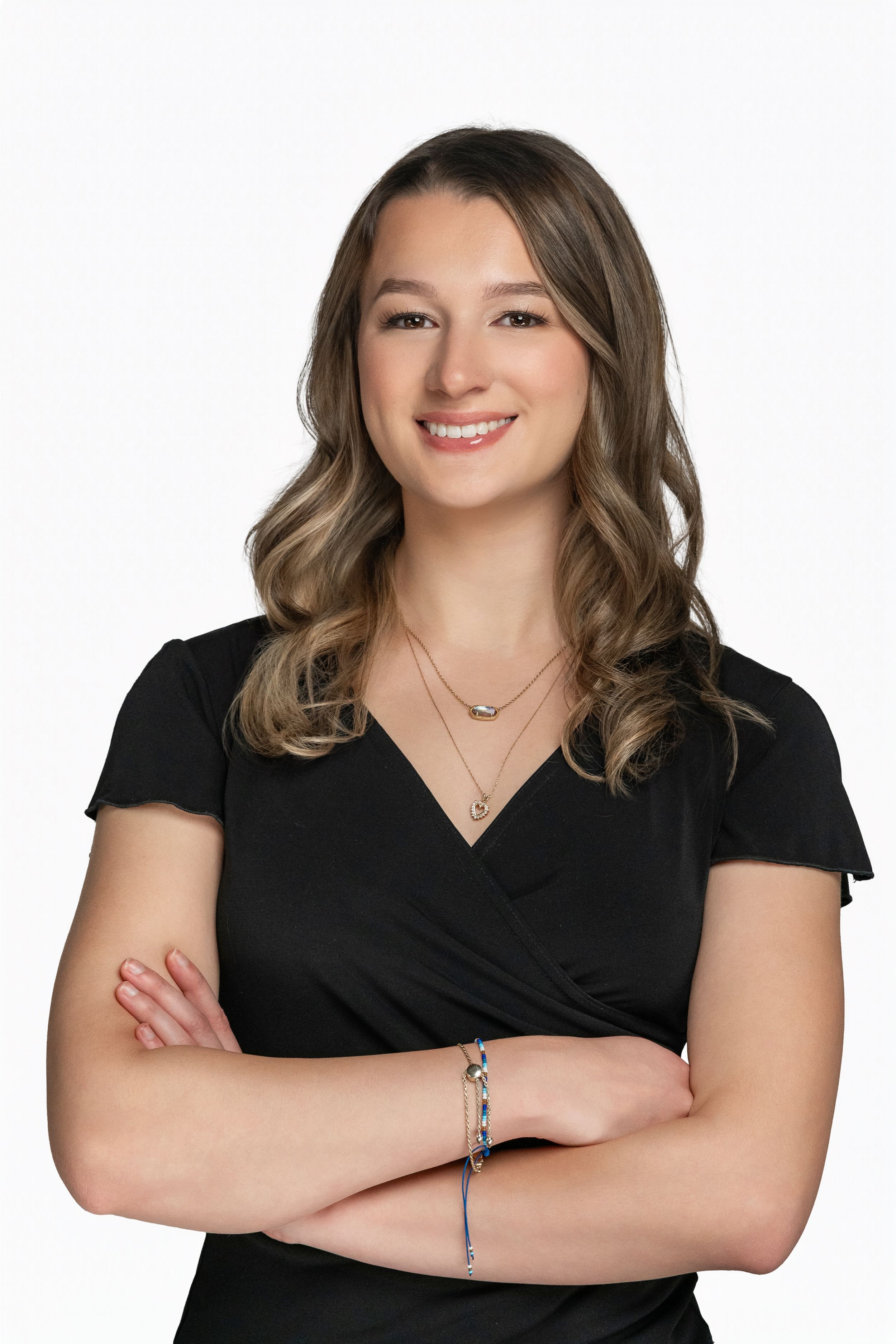 Professional headshot of someone in a black wrap dress with layered necklaces and wavy brown hair against white background.