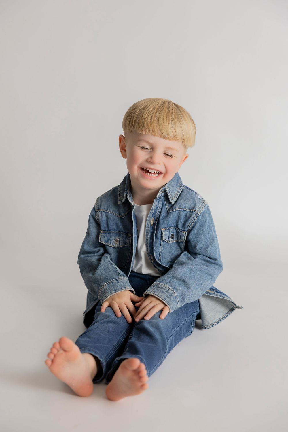A young child sits on a white backdrop wearing a denim jacket and jeans, smiling with bare feet at lpm collection studio in Pontiac michigan