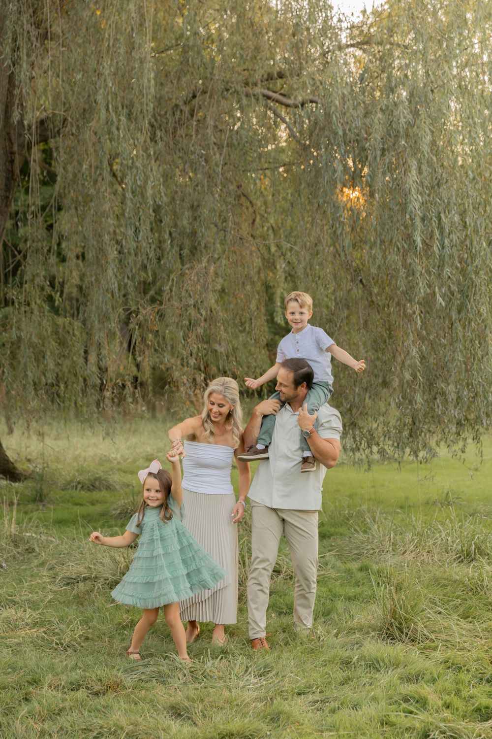 family of four playing with their children in a grassy field in front of a willow tree