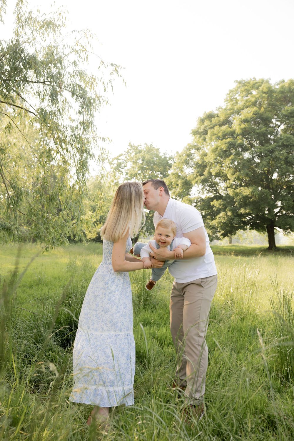 A family shares a tender moment together in a sunlit meadow with tall grass and willow trees in Lake Orion Michigan