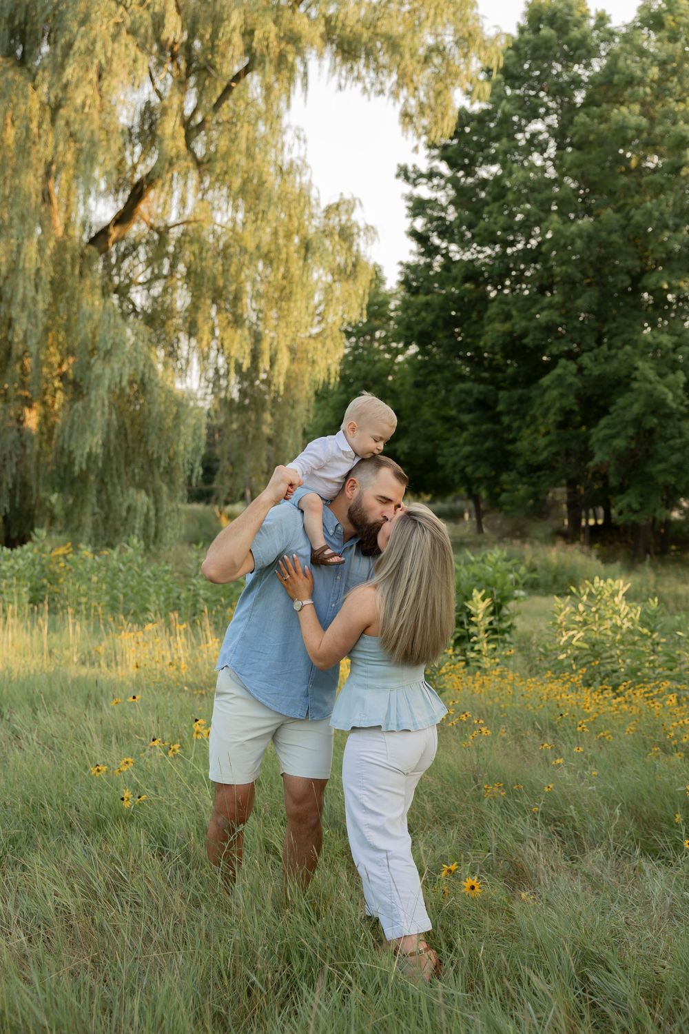 A couple plays with their baby on his shoulders while they kiss outdoors in a lush green meadow with wildflowers and willow trees.
