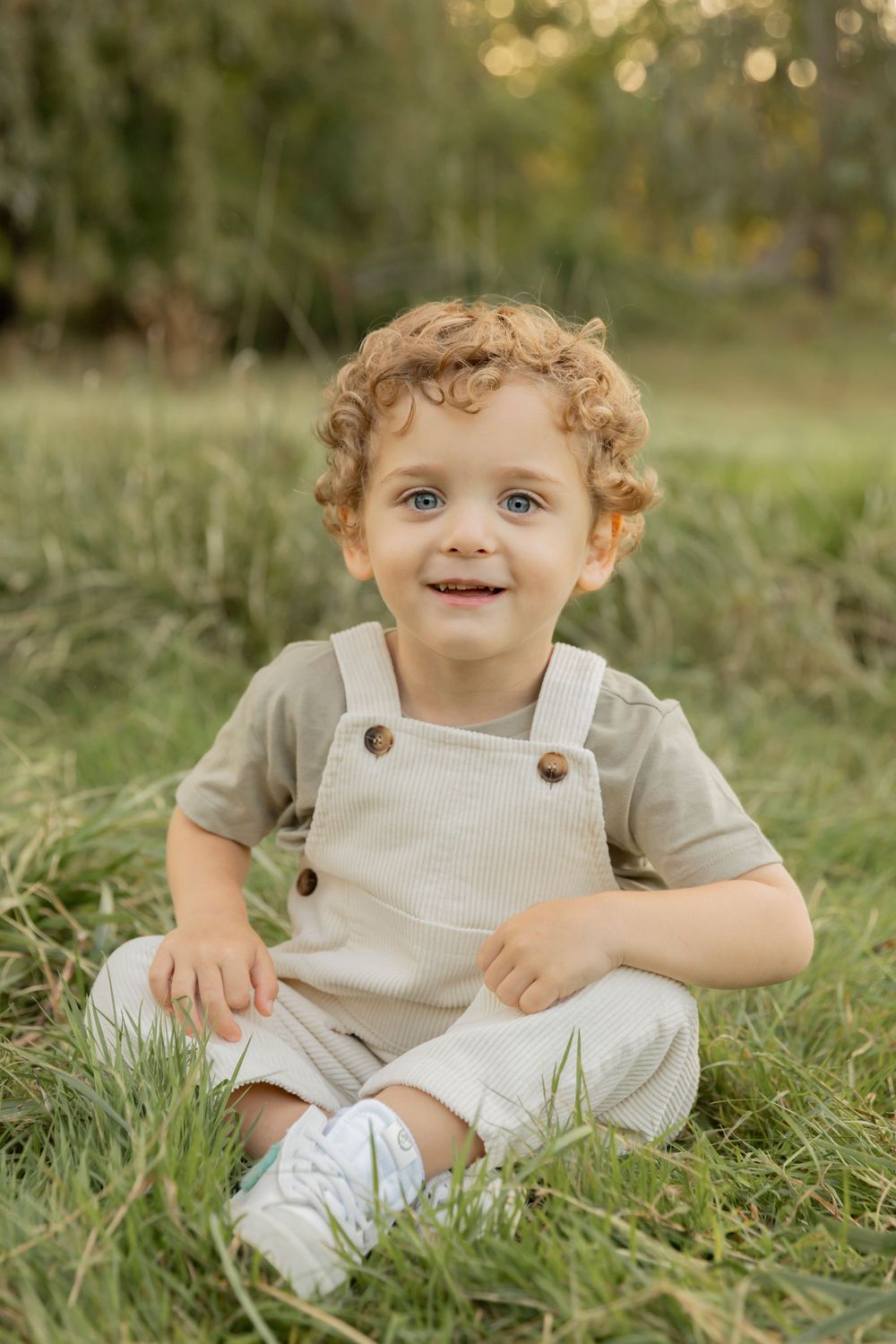 toddler boy in overalls sitting in a grassy field smiling for a photo