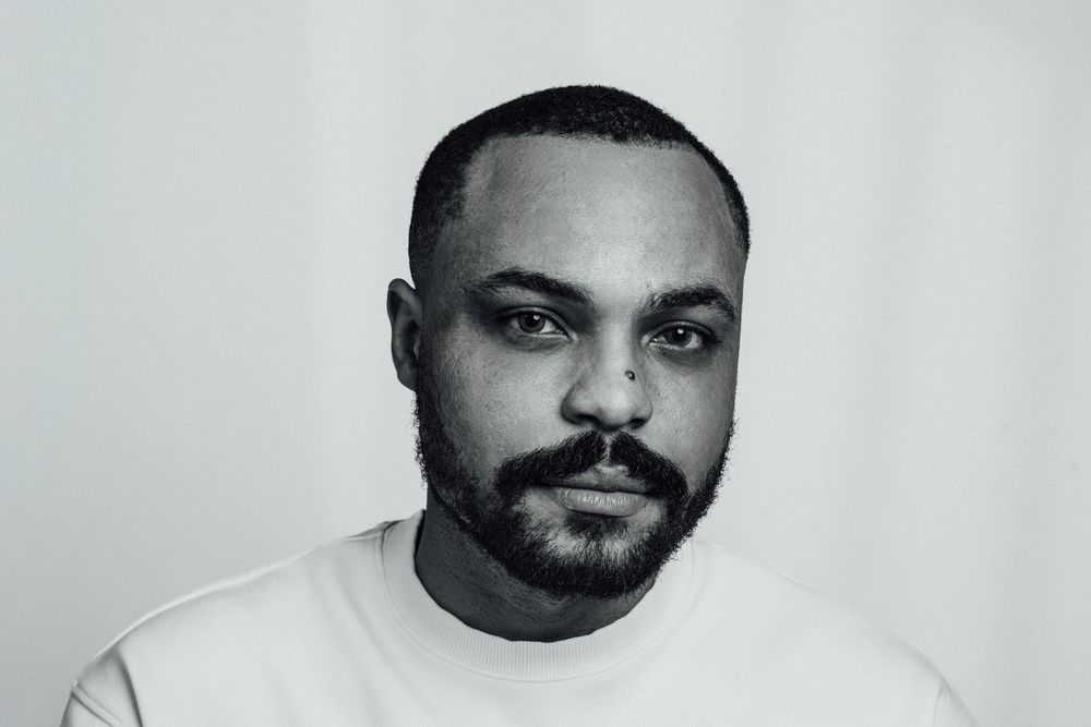 Black and white portrait of a young man with a beard wearing a white t-shirt against a light background.