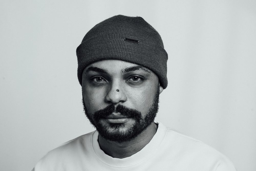 Black and white portrait of a bearded man wearing a gray beanie hat and white t-shirt against a light background.