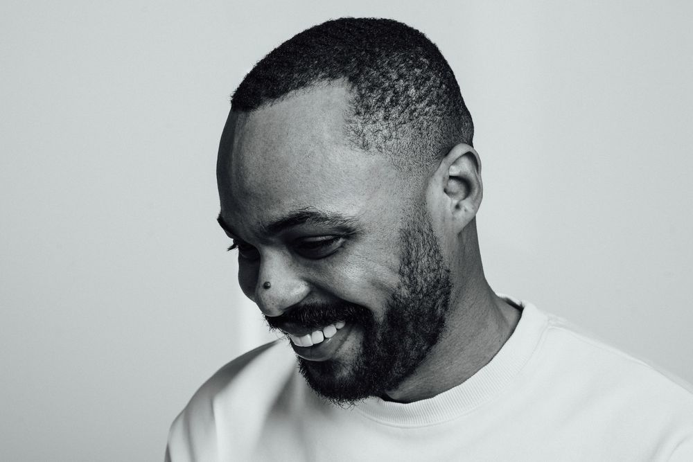 Black and white portrait of a smiling bearded Black man in a white t-shirt looking down.