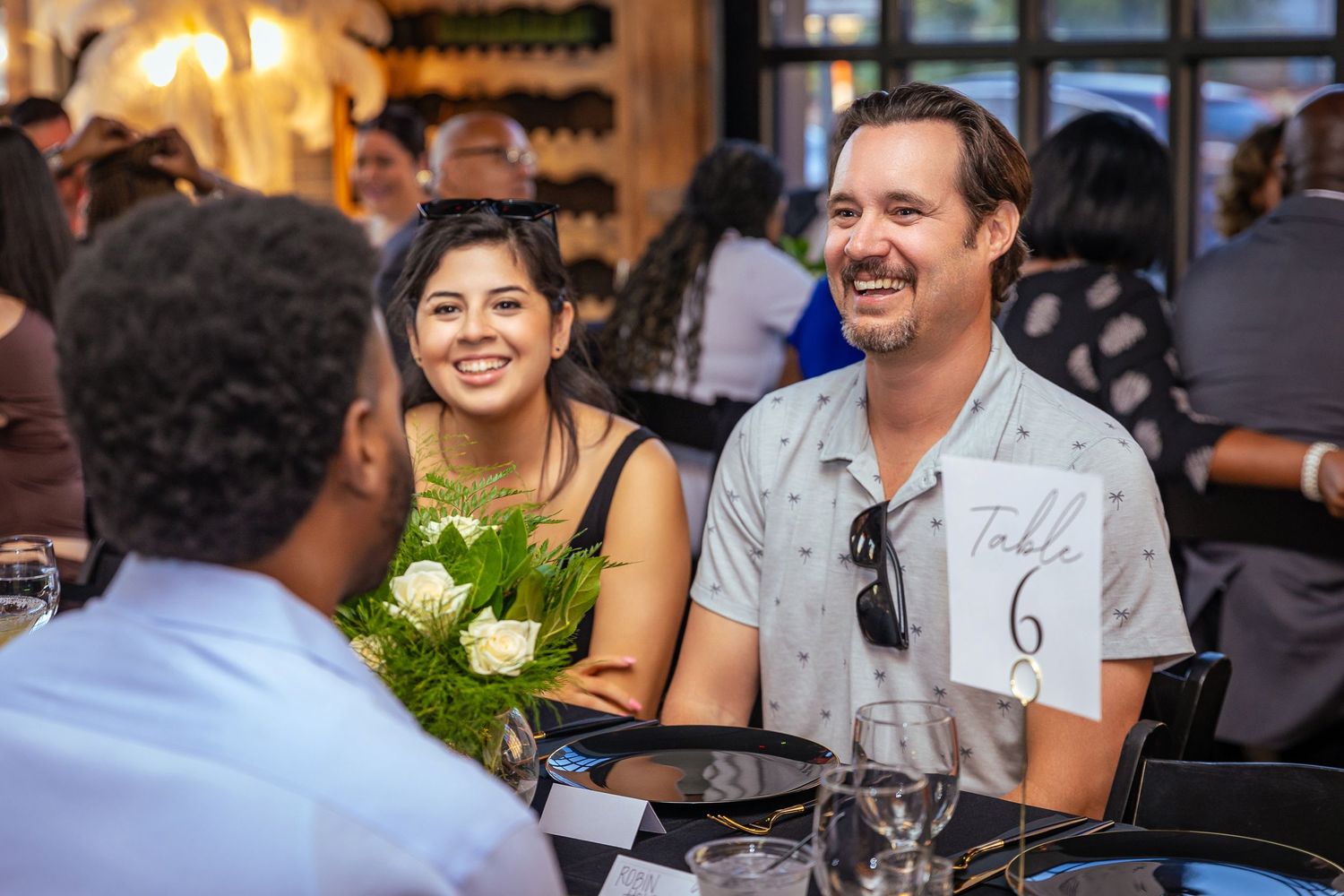 People laughing and enjoying dinner conversation at a restaurant table while one holds a flower bouquet.