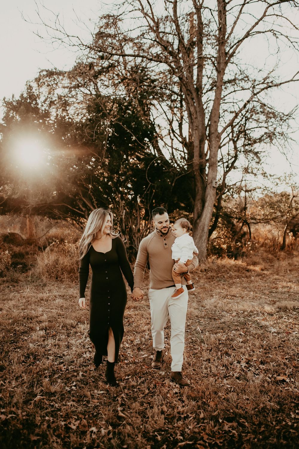 A couple walks through a sunlit autumn field with bare trees and fallen leaves in a warm sepia-toned photo series.