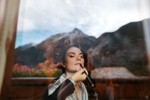 make up of the bride while standing near a window overlooking scenic mountains and autumn foliage.