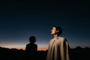 Two people stand outside silhouetted against a dark blue dusky sky with mountains in the background.