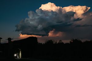 A dramatic sunset illuminates towering cumulonimbus clouds above a dark silhouetted landscape with a small light below.