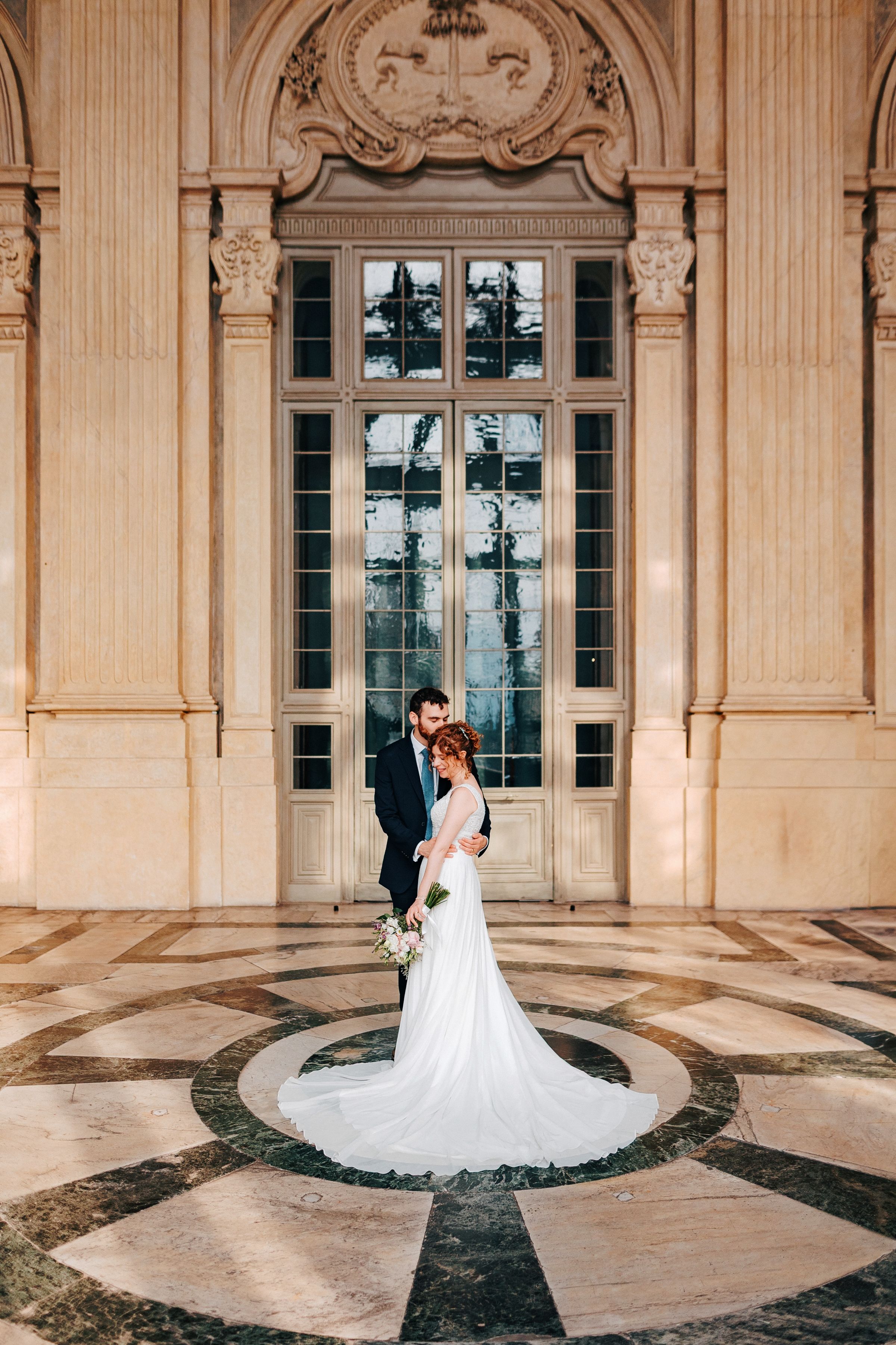 A wedding couple poses in front of an elegant stone archway with ornate architectural details and windows.