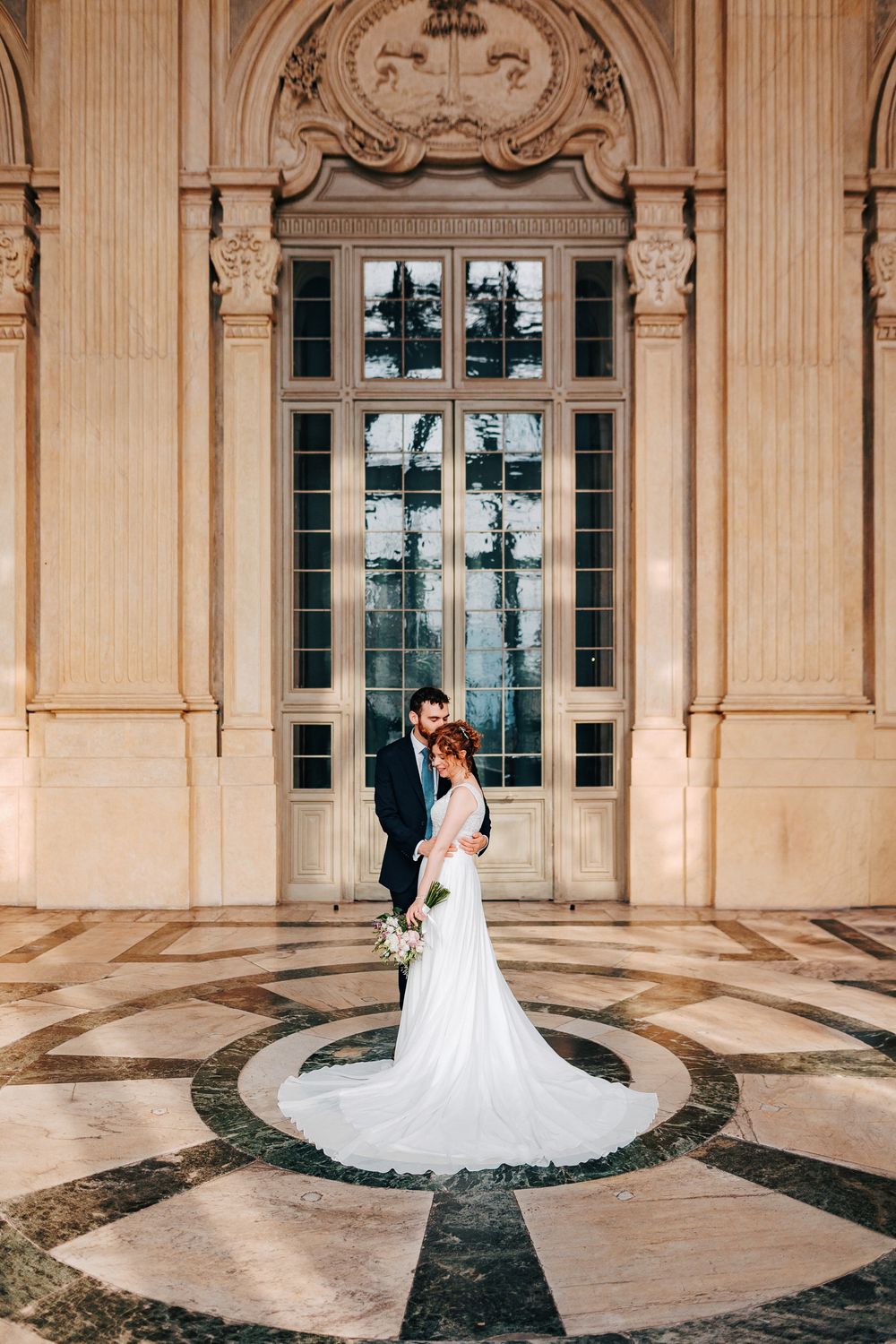 A wedding couple poses in front of an elegant stone archway with ornate architectural details and windows.