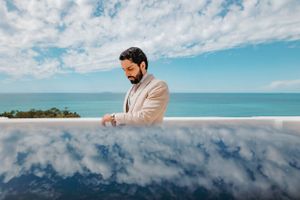 Reflection in infinity pool mirror shows person in beige blazer against tropical ocean and cloudy blue sky backdrop.
