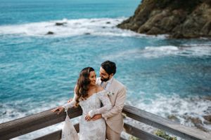 A romantic couple embraces on a coastal overlook with crashing turquoise waves and rocky cliffs in the background.