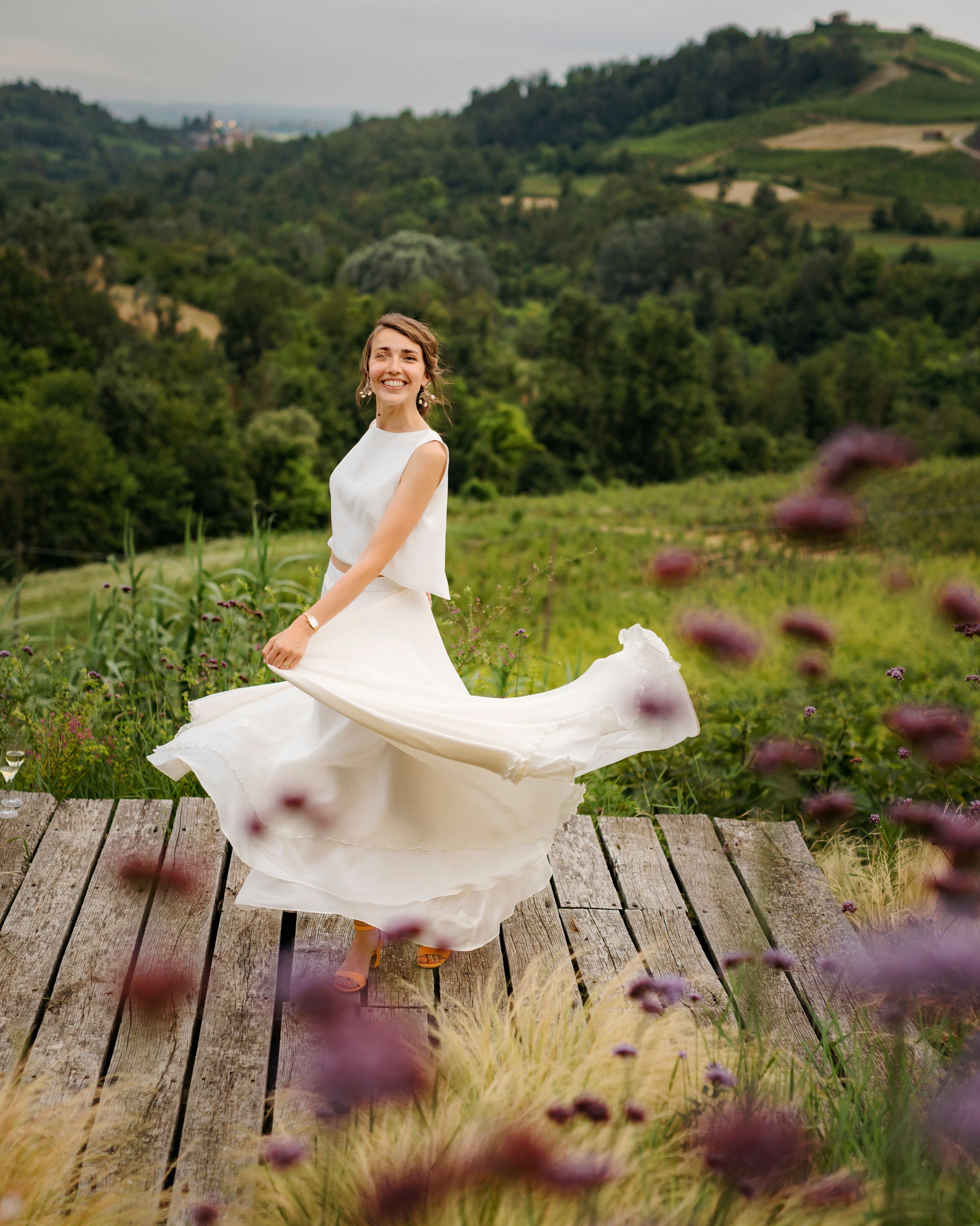 White dress flows in the wind on a wooden deck overlooking lush green mountain landscape with purple wildflowers.