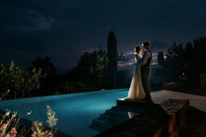A romantic couple embraces by an illuminated infinity pool at night with a dramatic moonlit sky in the background.