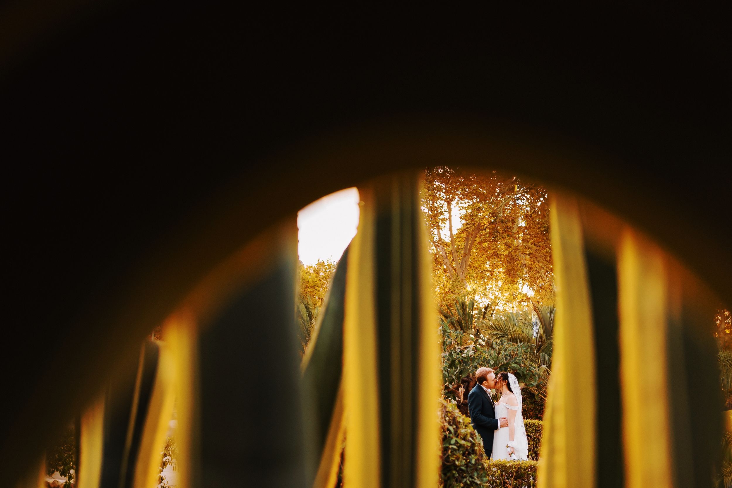 Creative wedding photo captured through golden railings showing couple in autumn garden setting.