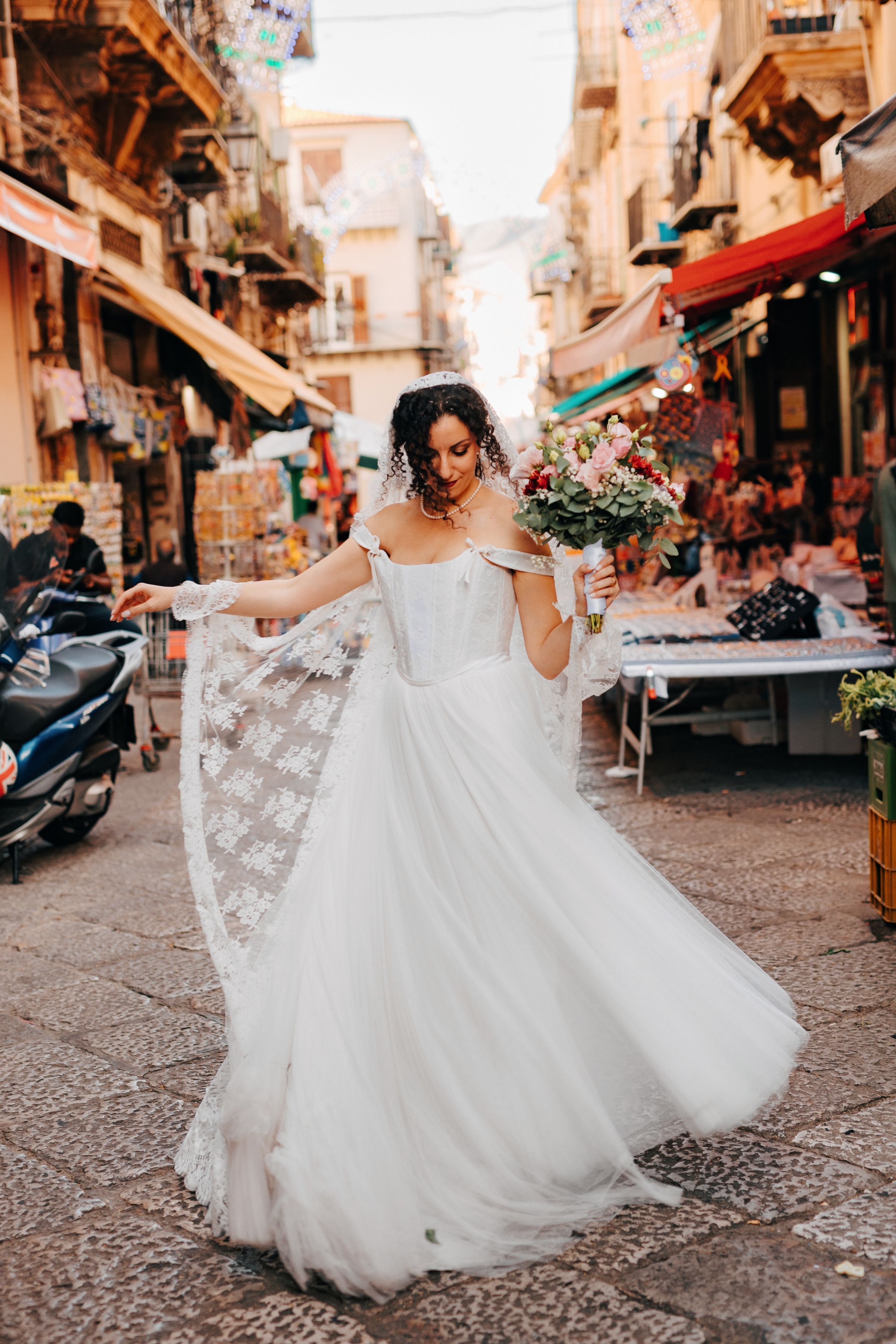 A bride in a flowing white gown twirls joyfully on a charming Italian street lined with shops and scooters.