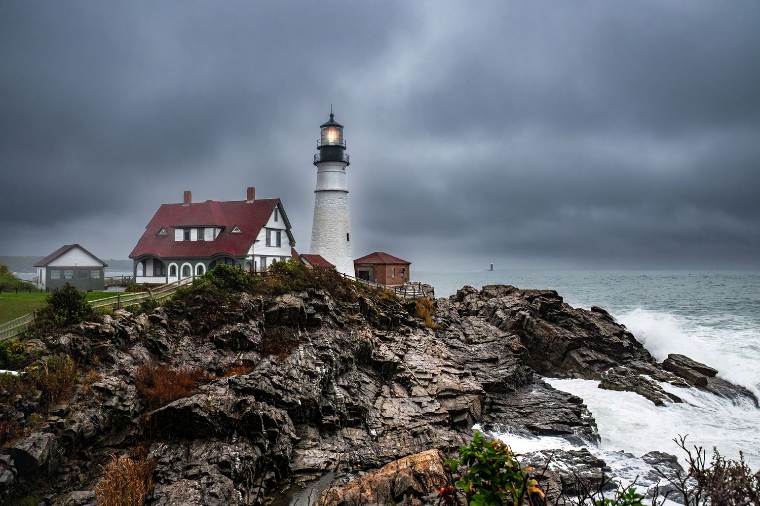 Dramatic coastal lighthouse perched on rocky cliffs with stormy waves crashing against New England shoreline.
