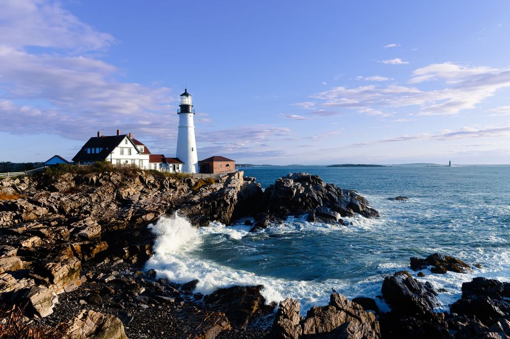 Waves crash against rocky cliffs near Portland Head Light lighthouse in Maine as ocean spray fills the air.