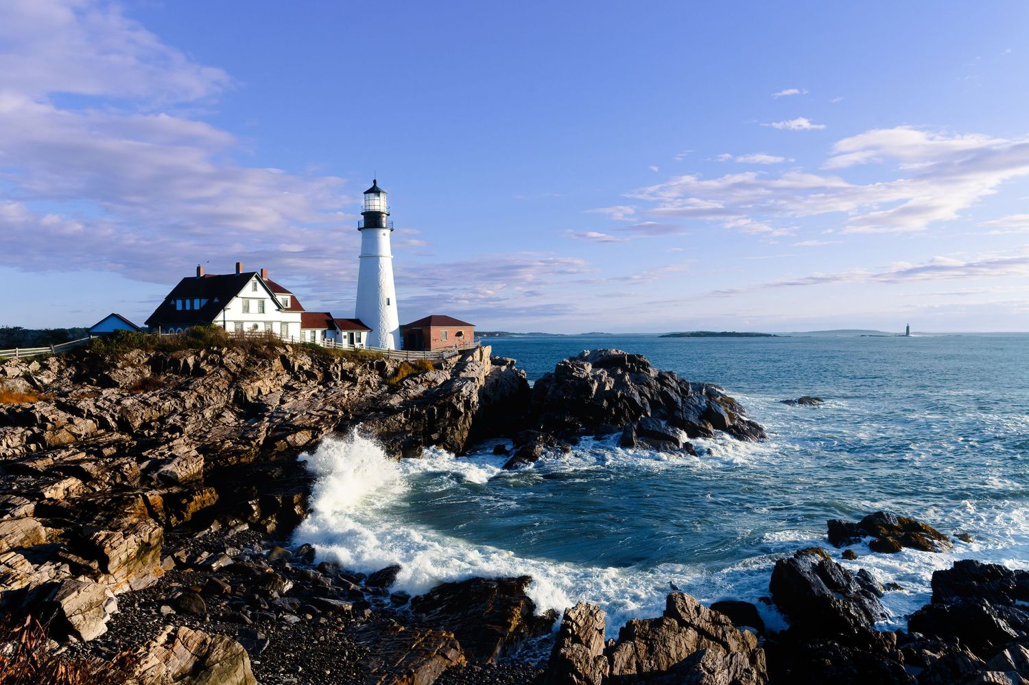 Waves crash against rocky cliffs near Portland Head Light lighthouse in Maine as ocean spray fills the air.