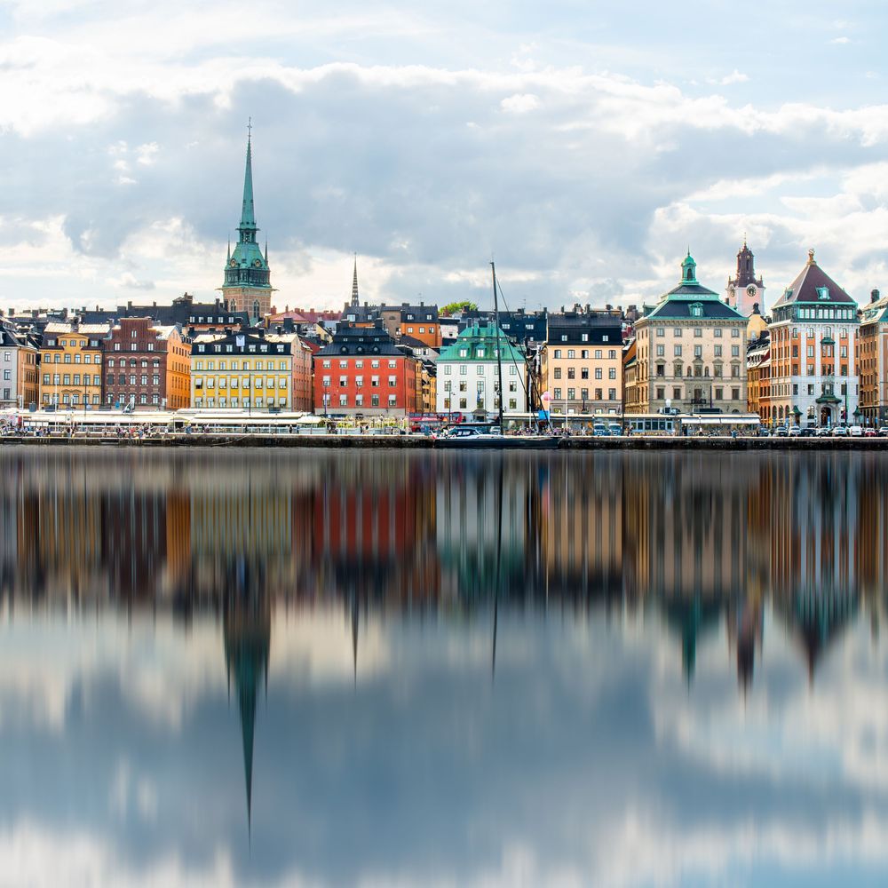 Colorful historic buildings reflected in calm waters along Stockholm's waterfront with church spires on the horizon.