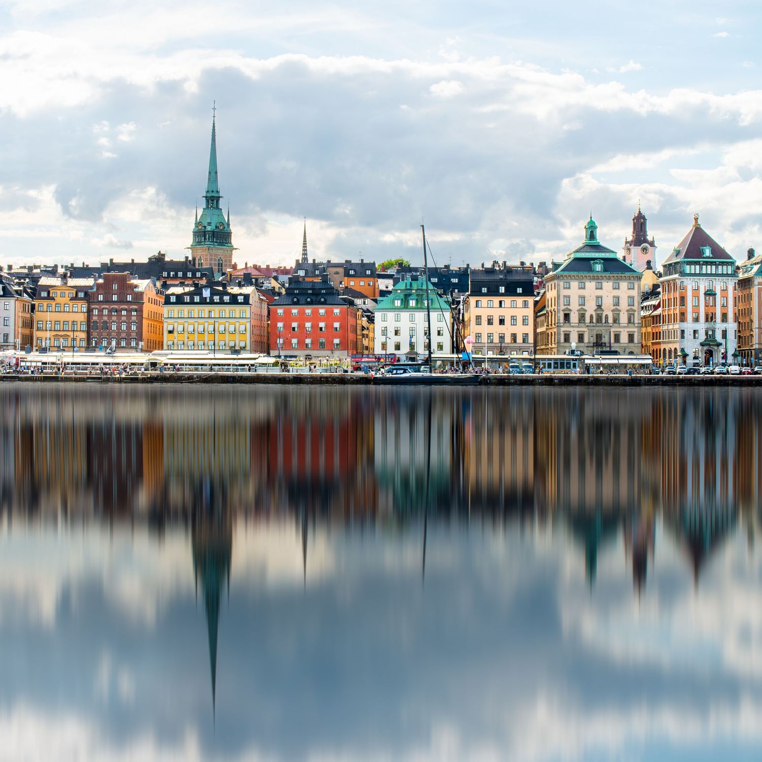 Colorful historic buildings reflected in calm waters along Stockholm's waterfront with church spires on the horizon.