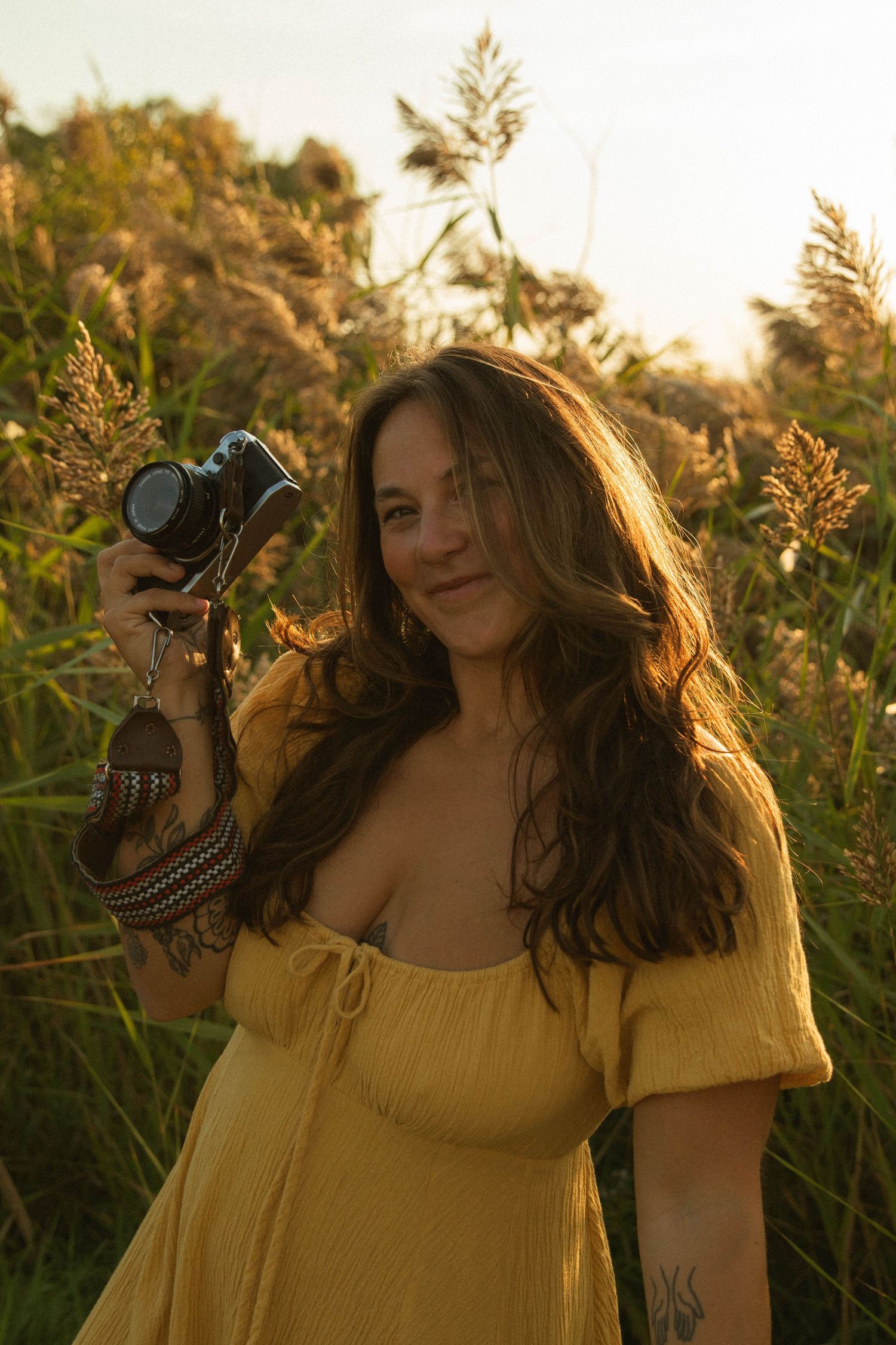 Young woman in yellow dress holding a film camera while standing in tall golden grass at sunset.
