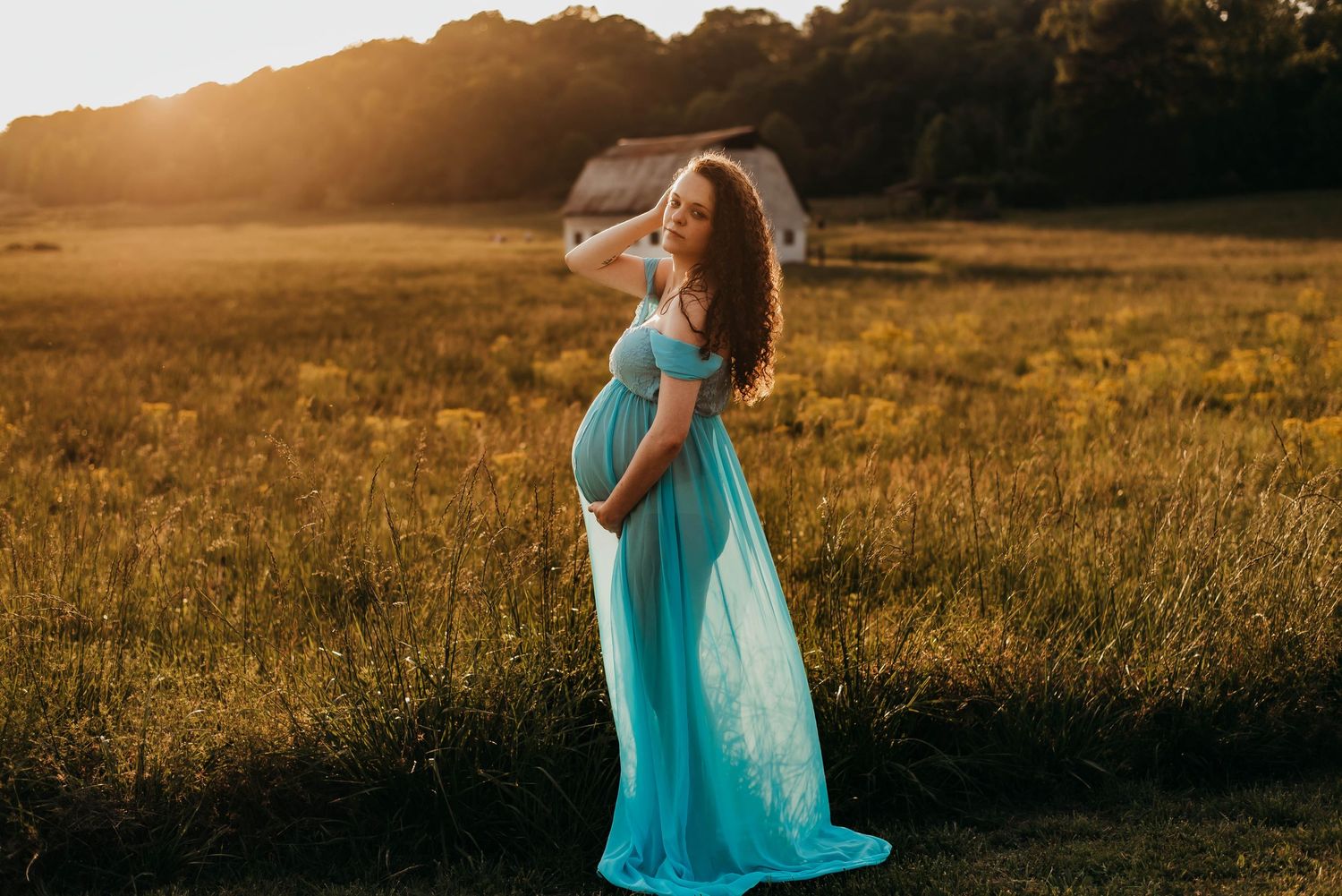 Pregnant woman in turquoise flowing maternity dress standing in sunlit golden field at sunset.