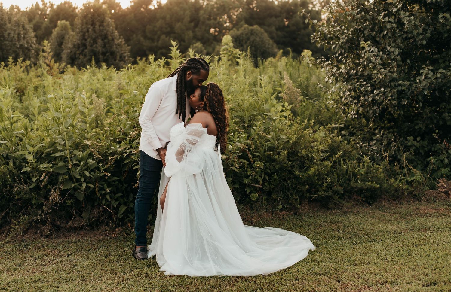 Couple sharing tender kiss outdoors, woman in flowing white maternity gown surrounded by lush greenery.
