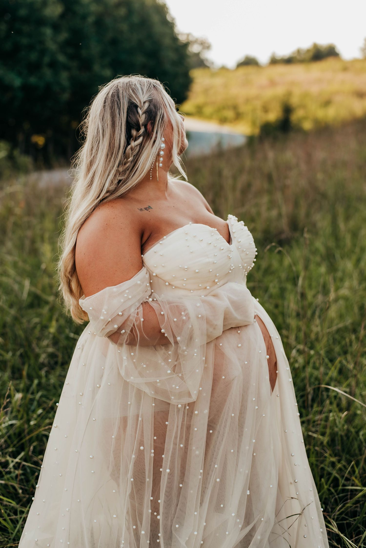 Pregnant woman in pearl-embellished off-shoulder tulle gown standing in golden meadow, back to camera.