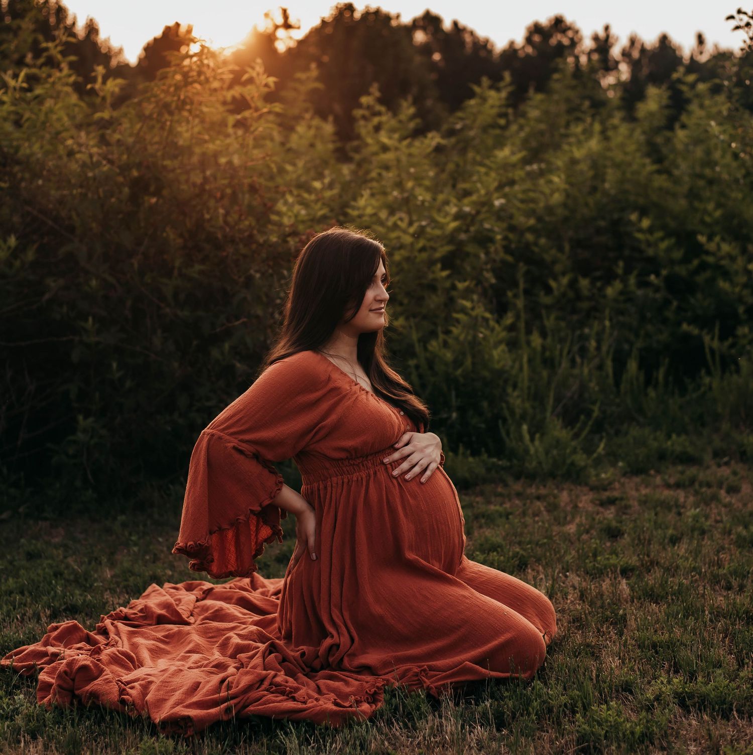Pregnant woman in rust orange dress sitting in grass during golden hour maternity photoshoot.