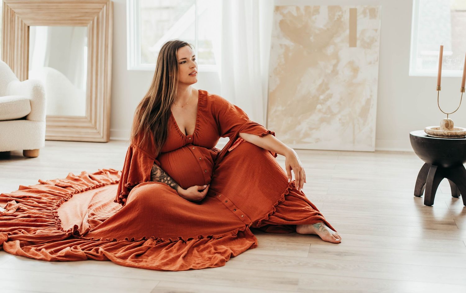 Pregnant woman in rust orange maternity gown sitting gracefully on floor during indoor photo session.
