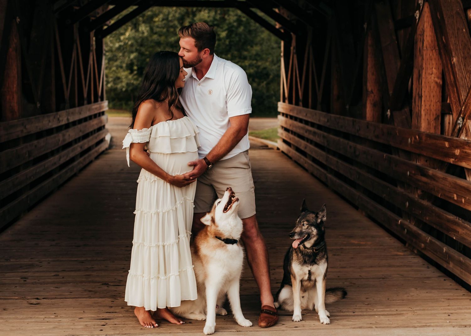 Expecting couple in white outfits posing with two dogs on rustic covered wooden bridge.